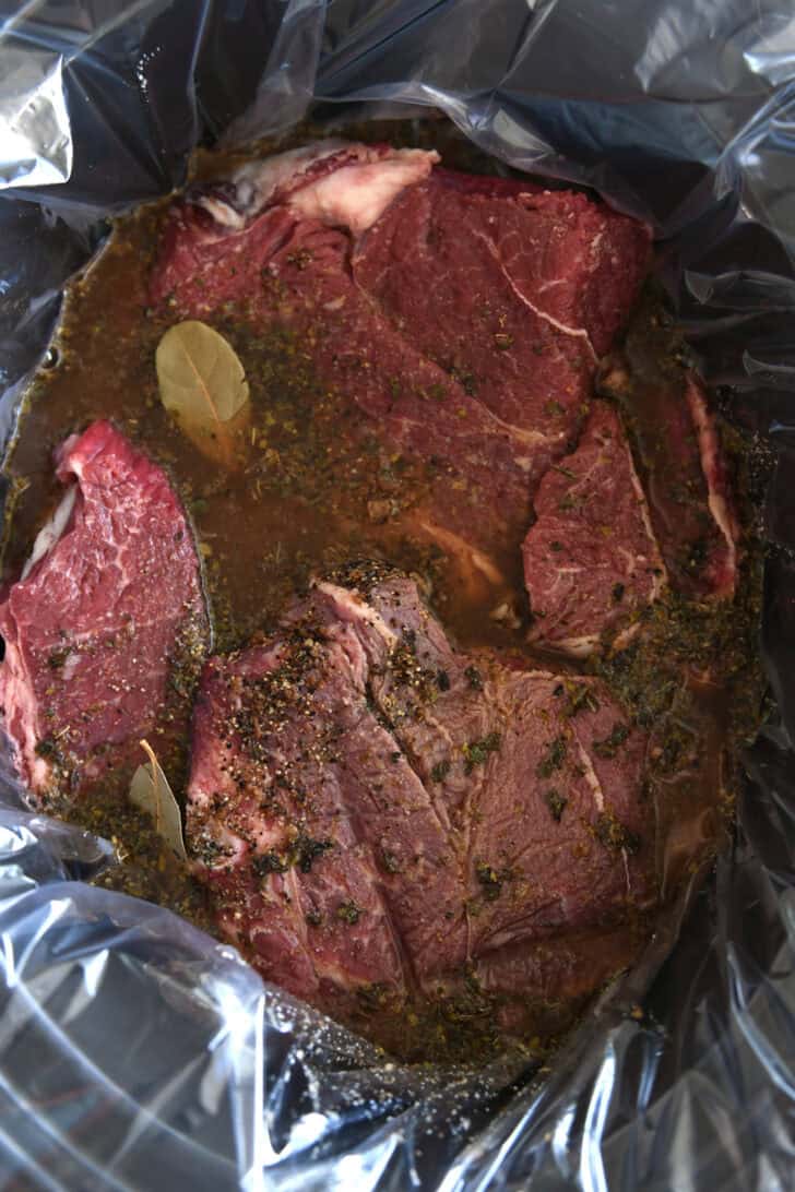 Black slow cooker lined with slow cooker liner and filled with chuck roast, bay leaves, and beef broth.