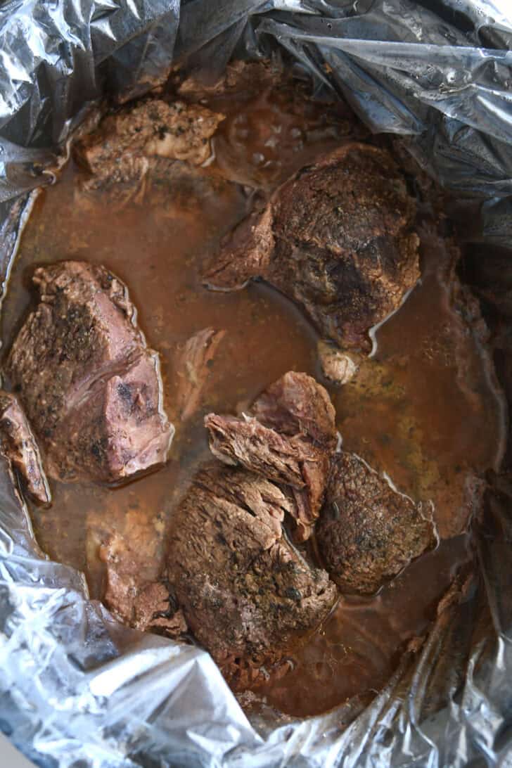 Black slow cooker insert with slow cooker liner filled with cooked beef and beef broth mixture.