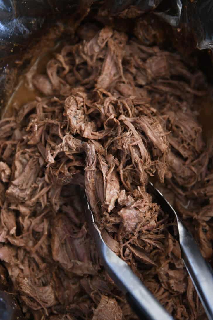 Tongs pinching serving of cooked, shredded Italian beef in black slow cooker liner.