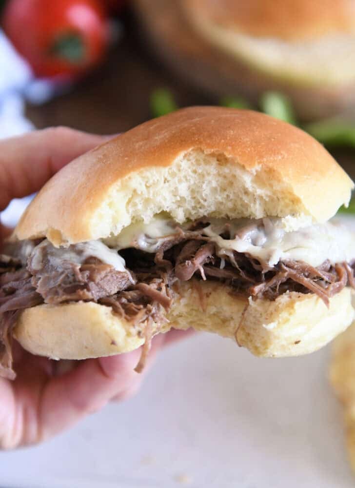 Slow cooker beef on crusty rolls with provolone cheese and one bite taken out; hand holding sandwich.