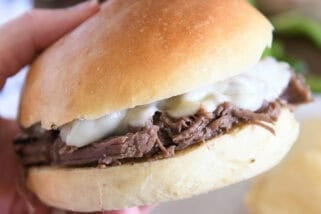 Slow cooker beef on crusty rolls with provolone cheese and one bite taken out; hand holding sandwich.