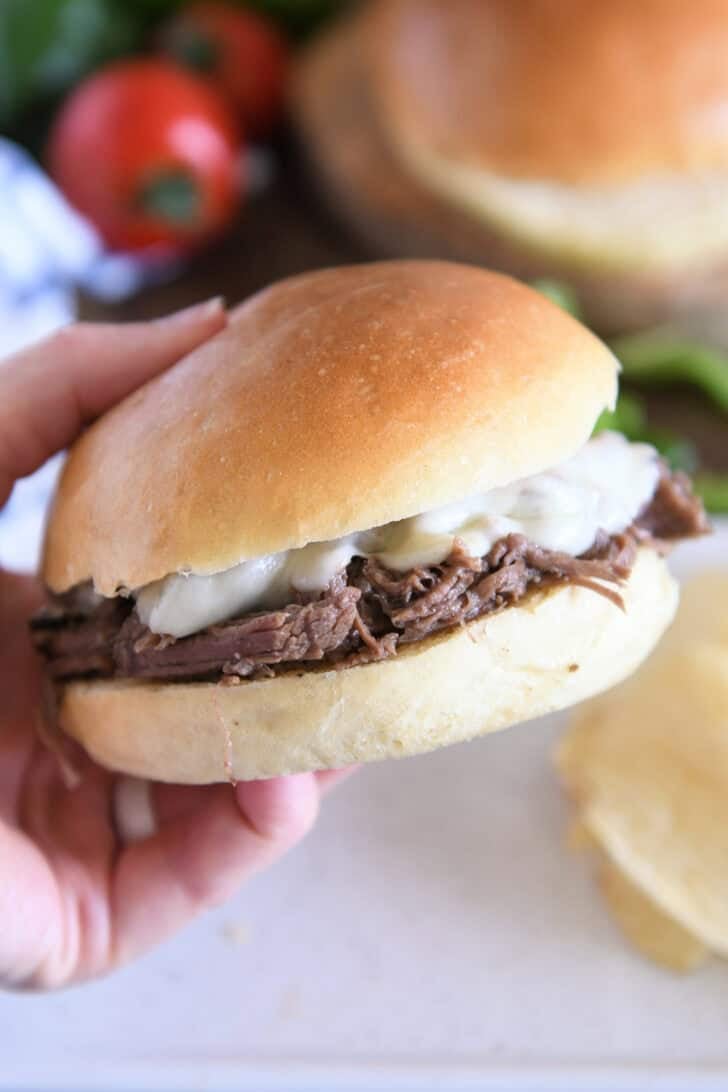 Slow cooker beef on crusty rolls with provolone cheese and one bite taken out; hand holding sandwich.