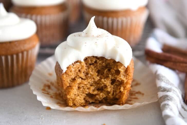 Pumpkin cupcake topped with whipped cream cheese frosting with bite taken out on unwrapped cupcake liner.