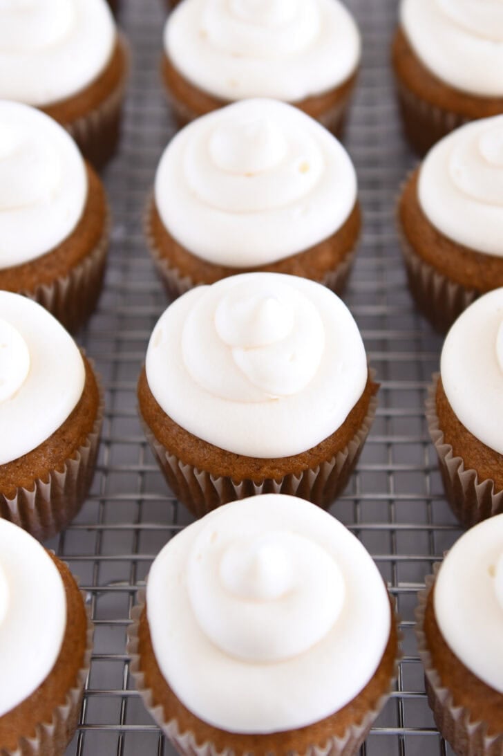 Lots of pumpkin cupcakes topped with cream cheese frosting on wire cooling rack.