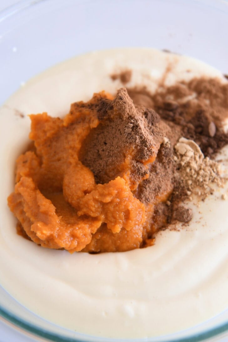 Cheesecake batter in glass bowl with canned pumpkin, cinnamon, and nutmeg.