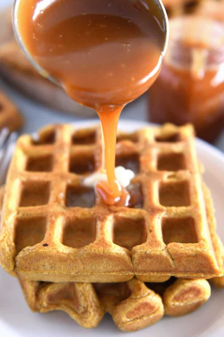 Small ladle drizzling caramel syrup over pumpkin waffles on white plate.