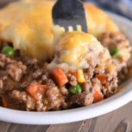 A beef and vegetable mixture topped with cheesy potatoes in a white bowl. A fork is taking a bite out.