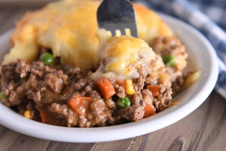 A beef and vegetable mixture topped with cheesy potatoes in a white bowl. A fork is taking a bite out.