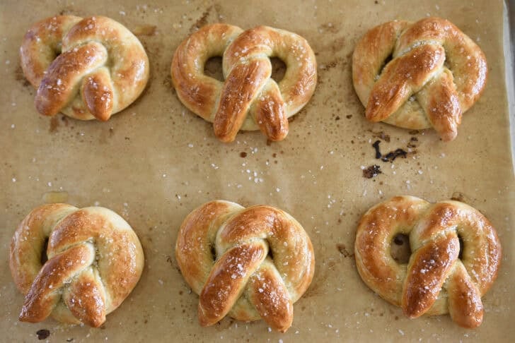 Six home-baked soft pretzels on parchment-lined baking sheet and sprinkled with salt.