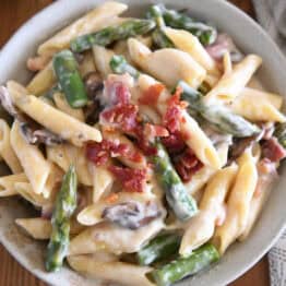 Top down view of creamy penne pasta with mushrooms, asparagus, and crumbled bacon in a white bowl.