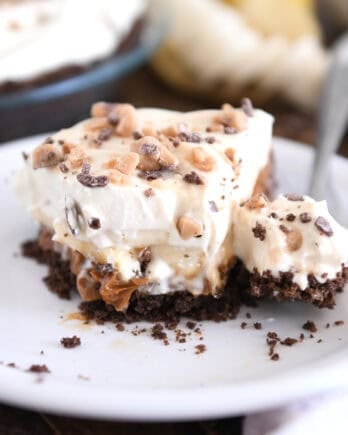 Slice of pie with chocolate cookie crust, dulce de leche, bananas, whipped cream and toffee on white plate.