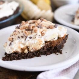 Slice of pie with chocolate cookie crust, dulce de leche, bananas, whipped cream and toffee on white plate.