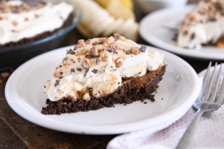 Slice of pie with chocolate cookie crust, dulce de leche, bananas, whipped cream and toffee on white plate.