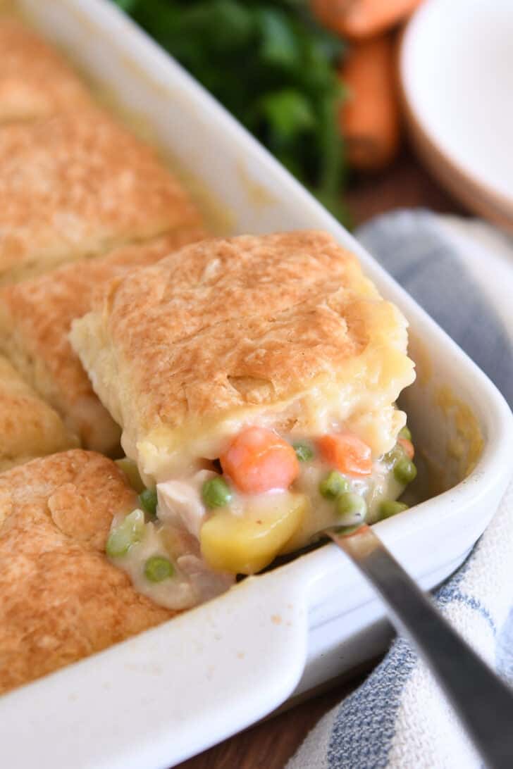 Spoon lifting out scoop of chicken pot pie filing for white 9X13-inch pan with baked biscuit on top.