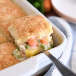 Spoon lifting out scoop of chicken pot pie filing for white 9X13-inch pan with baked biscuit on top.