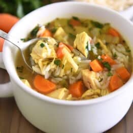 Spoon lifting up scoop of chicken and rice soup in white bowl with handles topped with fresh parsley and Parmesan cheese.