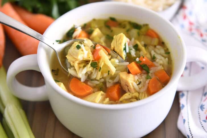 Spoon lifting up scoop of chicken and rice soup in white bowl with handles topped with fresh parsley and Parmesan cheese.