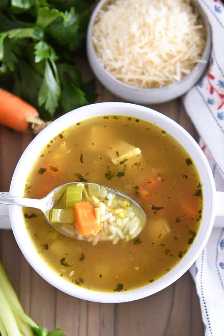 Spoon lifting up scoop of chicken and rice soup in white bowl with handles topped with fresh parsley and Parmesan cheese.
