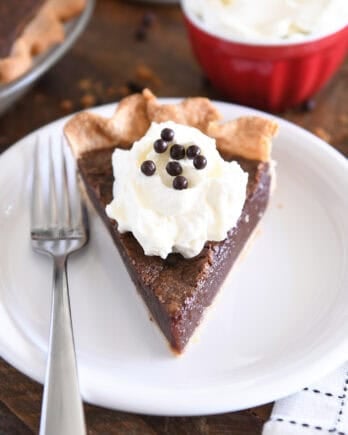 Triangle slice of chocolate chess pie on white plate with fork to the side and a dollop of whipped cream on top of pie topped with chocolate crisp pearls.