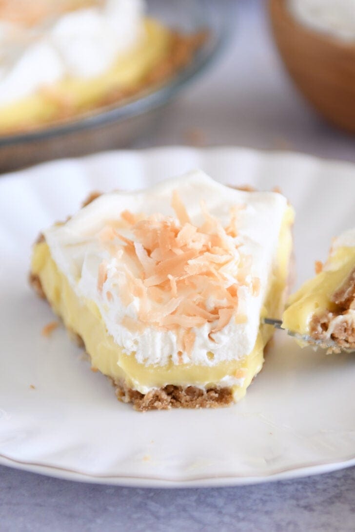 Slice of coconut cream pie topped with whipped cream and toasted coconut on white plate with one bite removed.