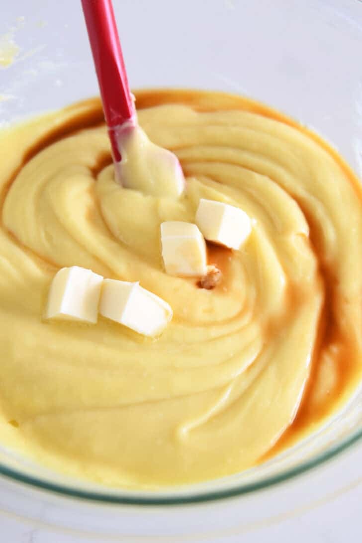 Several cubes butter in warm pudding with vanilla and spatula in glass bowl.