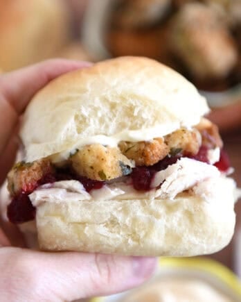 Holding homemade roll sandwiched with mayonnaise, stuffing, cranberry sauce and shredded turkey between fingers.