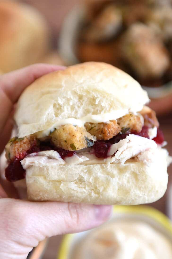 Holding homemade roll sandwiched with mayonnaise, stuffing, cranberry sauce and shredded turkey between fingers.