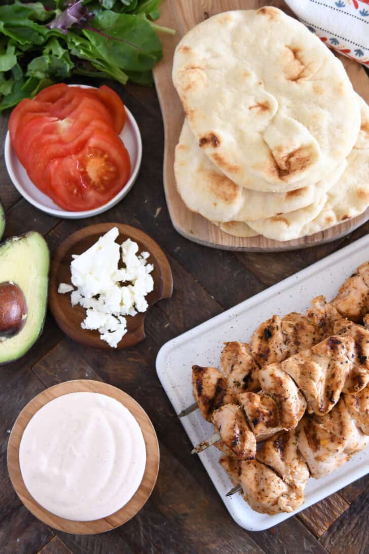Top down view of grilled chicken kebabs on a white tray, with a white sauce in a bowl, a stack of flatbread, and toppings around the kebabs.