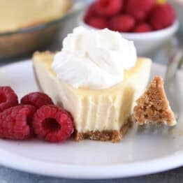 A slice of lemon pie with a graham cracker crust, topped with whipped cream and a bite taken out. Fresh raspberries sit beside the slice.