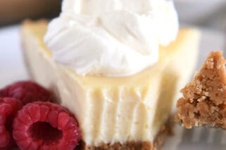 A slice of lemon pie with a graham cracker crust, topped with whipped cream and a bite taken out. Fresh raspberries sit beside the slice.