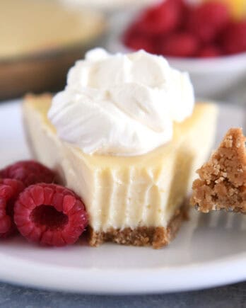 A slice of lemon pie with a graham cracker crust, topped with whipped cream and a bite taken out. Fresh raspberries sit beside the slice.