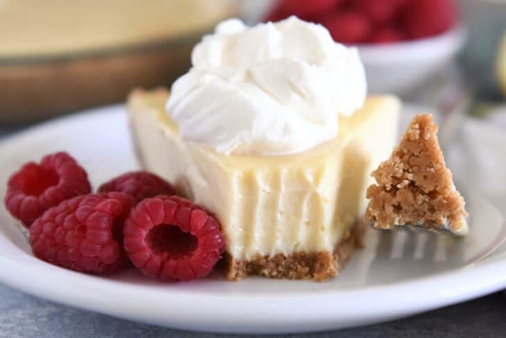 A slice of lemon pie with a graham cracker crust, topped with whipped cream and a bite taken out. Fresh raspberries sit beside the slice.