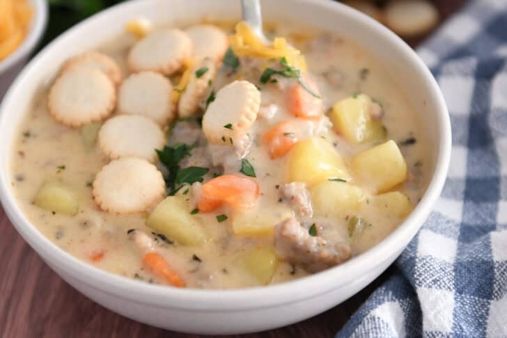 Soup bowl with sausage potato soup topped with oyster saltine crackers, fresh parsley and grated cheese and spoon on side.