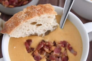Top down view of butternut squash soup in white bowl topped with bacon and piece of crusty bread.