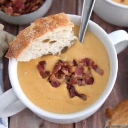 Top down view of butternut squash soup in white bowl with piece of crusty bread and cooked bacon pieces.