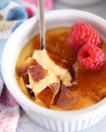 Spoon cracking through top shell of creme brûlée in white ramekin with raspberries on the side.