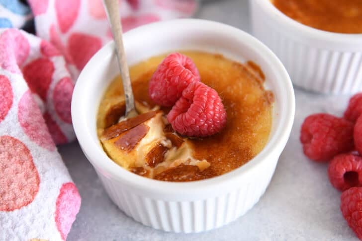 Spoon cracking through top shell of creme brûlée in white ramekin with raspberries on the side.