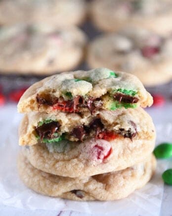 Two baked cookies with chocolate chips and red and green M&Ms with a cookie on top broken in half with melted chocolate chips.