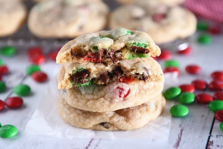 Two baked cookies with chocolate chips and red and green M&Ms with a cookie on top broken in half with melted chocolate chips.