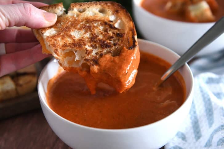White bowl with homemade tomato soup and half a grilled cheese dipped in the soup.