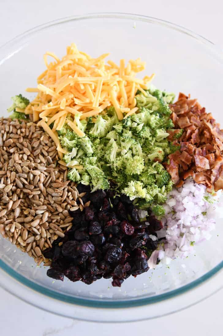 Top down view of a glass bowl with shredded cheese, sunflower seeds, chopped broccoli, bacon bits, dried cranberries, and diced onions.