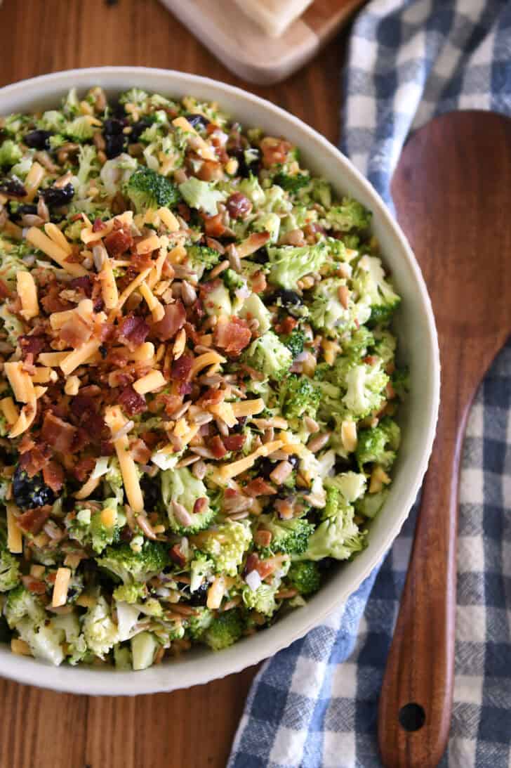 Top down view of a white bowl of chopped broccoli salad with shredded cheese, diced onion, bacon bits, sunflower seeds, and dried cranberries coated in a light sauce.