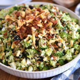 A white bowl of chopped broccoli salad with shredded cheese, diced onion, bacon bits, sunflower seeds, and dried cranberries coated in a light sauce.