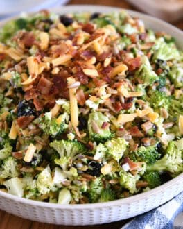 A white bowl of chopped broccoli salad with shredded cheese, diced onion, bacon bits, sunflower seeds, and dried cranberries coated in a light sauce.