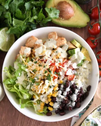 White bowl with lettuce, roasted corn, shredded cheese, tomatoes, black beans, avocados, cilantro lime rice and chicken.