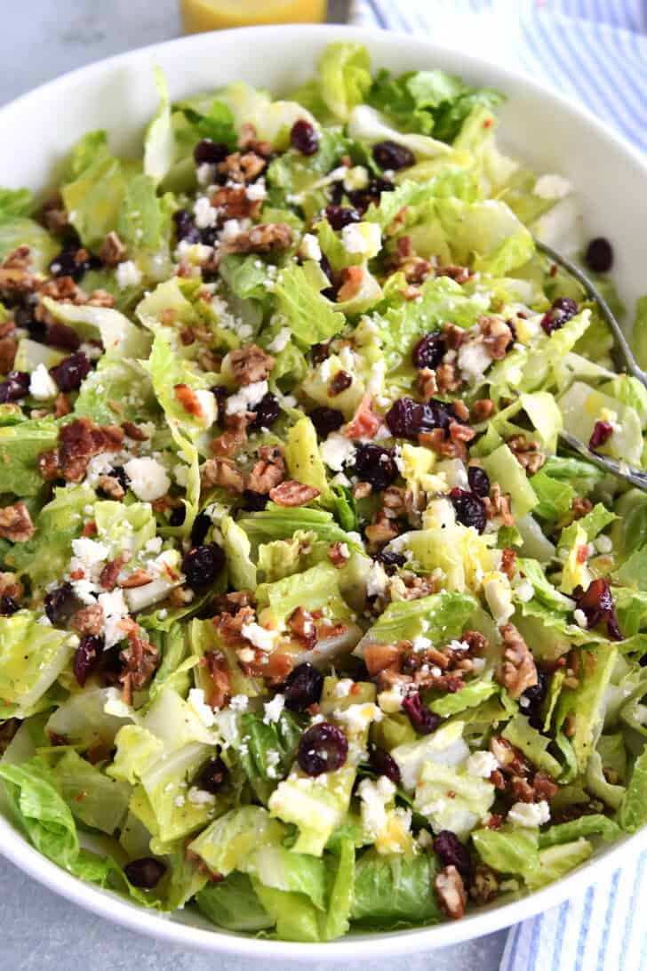 Top down view of a large white bowl full of chopped romaine salad with candied nuts, bacon bits, crumbled cheese, and dried cranberries.