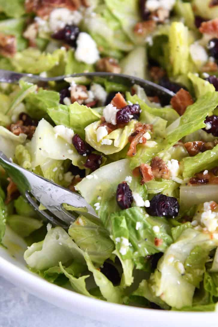 Metal tongs getting out a serving of chopped romaine salad with bacon bits, crumbled cheese, and dried cranberries from a white bowl.