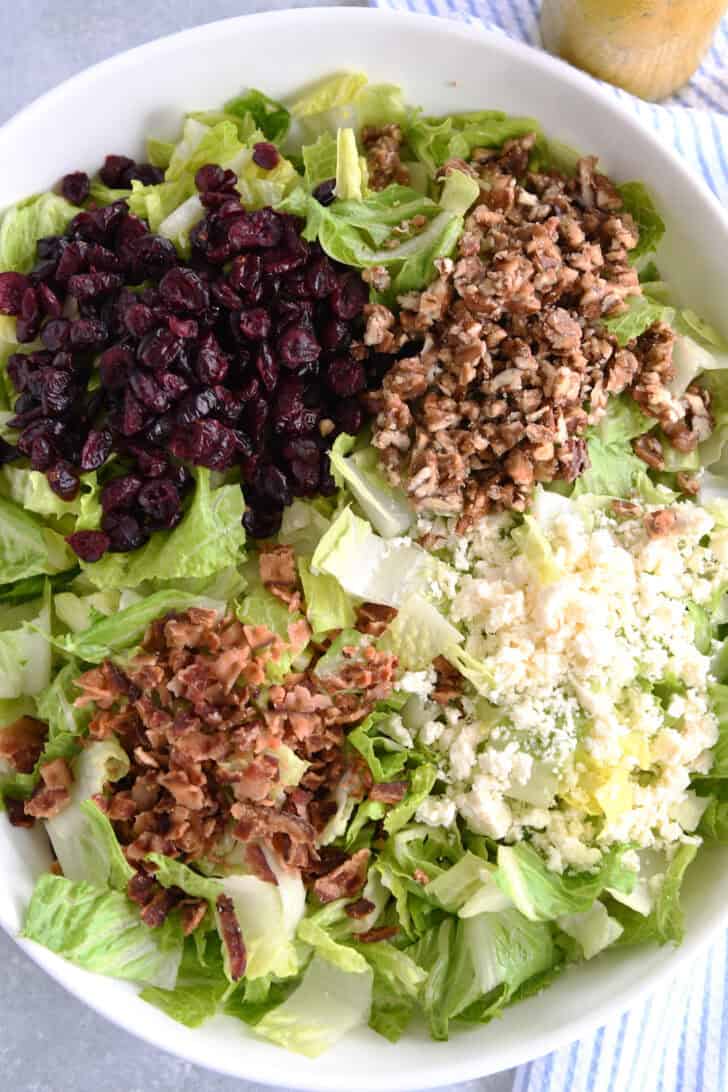 Top down view of an oval white bowl with chopped romaine salad, chopped nuts, crumbled cheese, dried cranberries, and bacon bits separated into piles on top of the lettuce.
