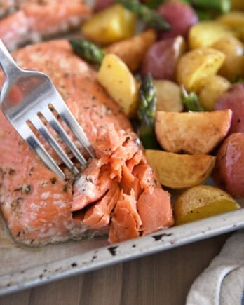 Fork flaking filet of baked salmon with roasted potatoes and asparagus on sheet pan.