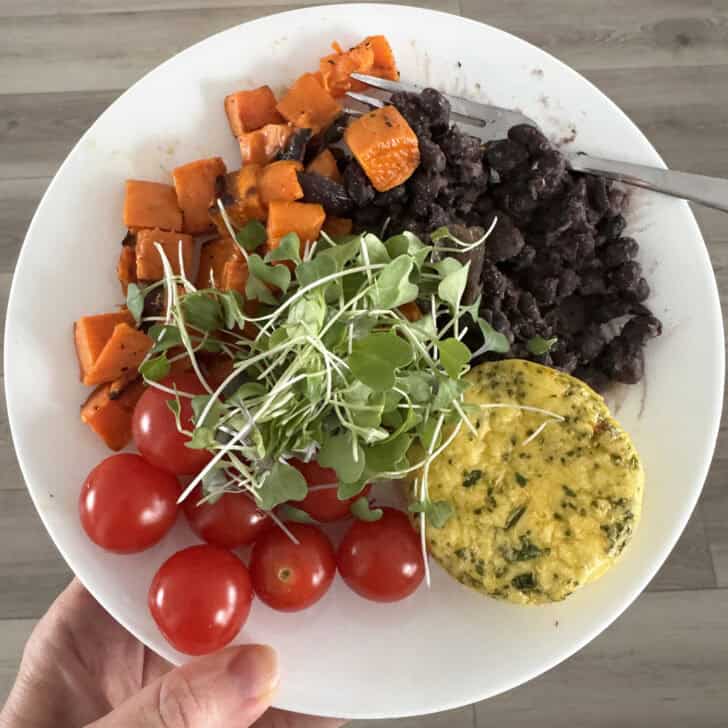 White plate with egg bite, black beans, sweet potatoes, cherry tomatoes and microgreens.